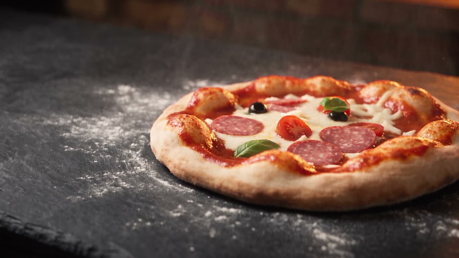 Pizza mit geschmolzenem Käse und Basilikum auf dunklem Schiefer, im Hintergrund die Glut des Steinofens
